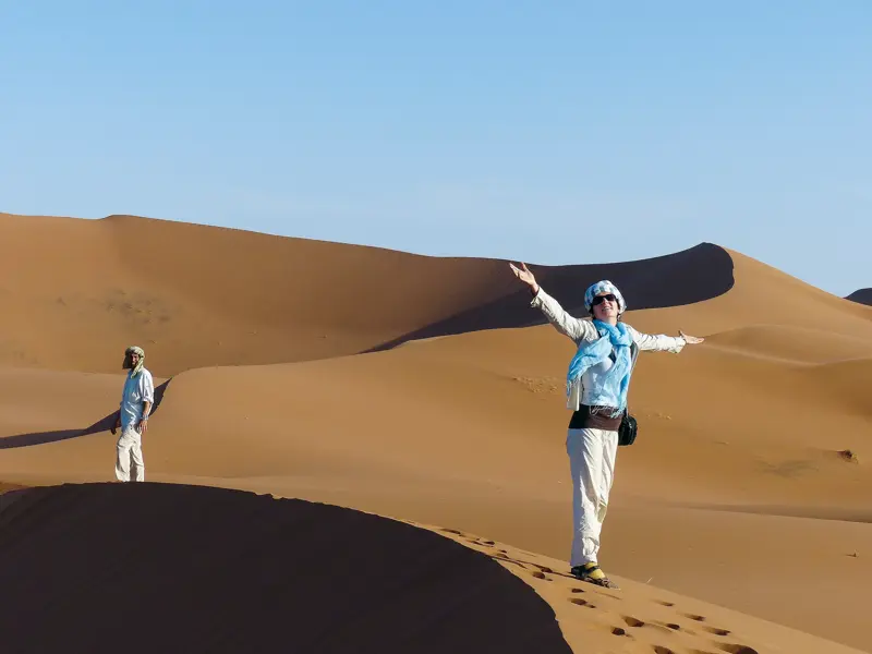 Glückliche Wanderin mit offenen Armen auf einer Sanddüne beim Wüstentrekking im Erg Chebbi, Marokko, mit lokalem Guide.