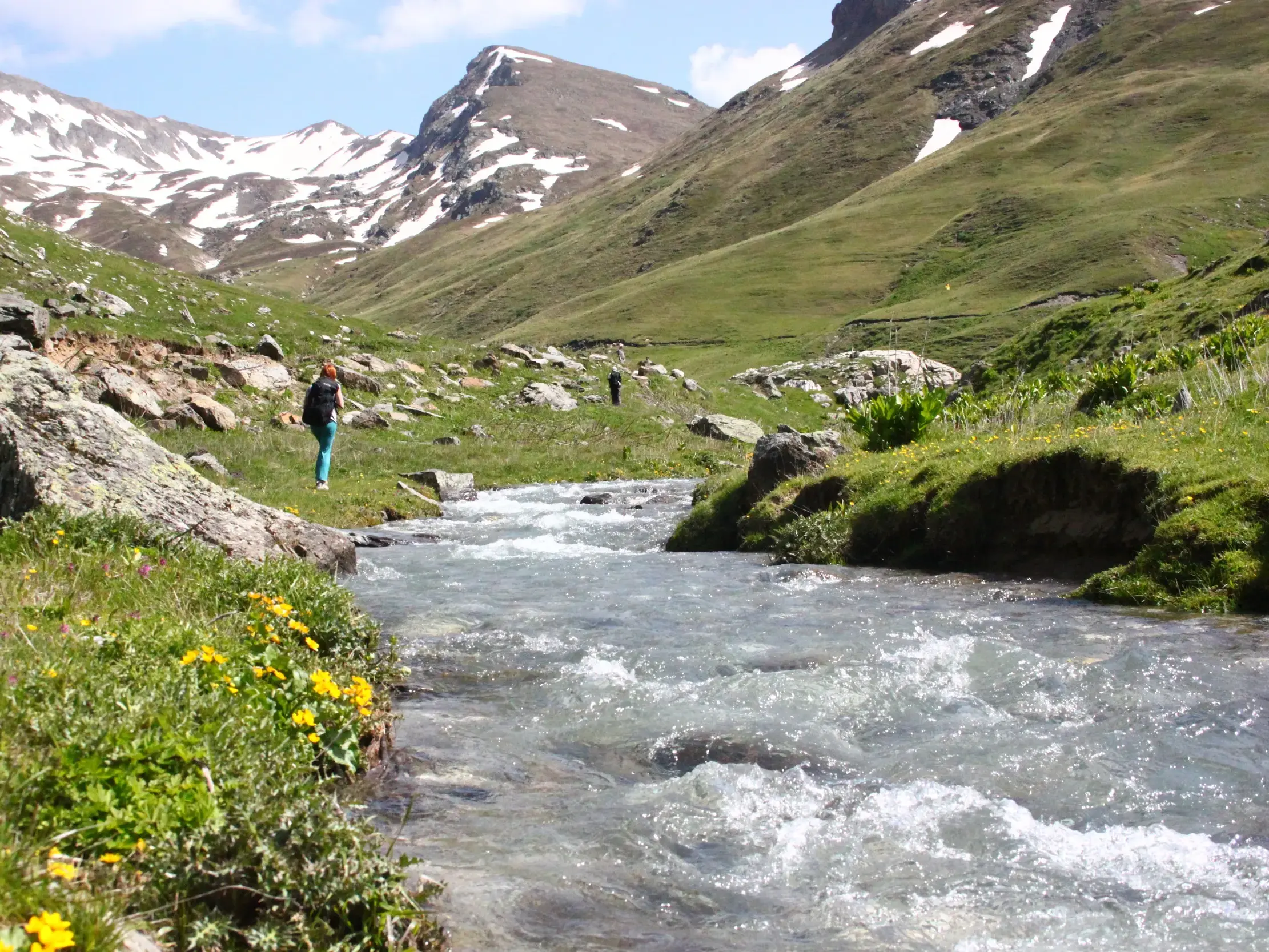 Eine Wanderin pausiert am Ufer eines Gebirgsflusses im Korab-Koritnik Naturpark vor schneebedeckten Gipfeln in Albanien.