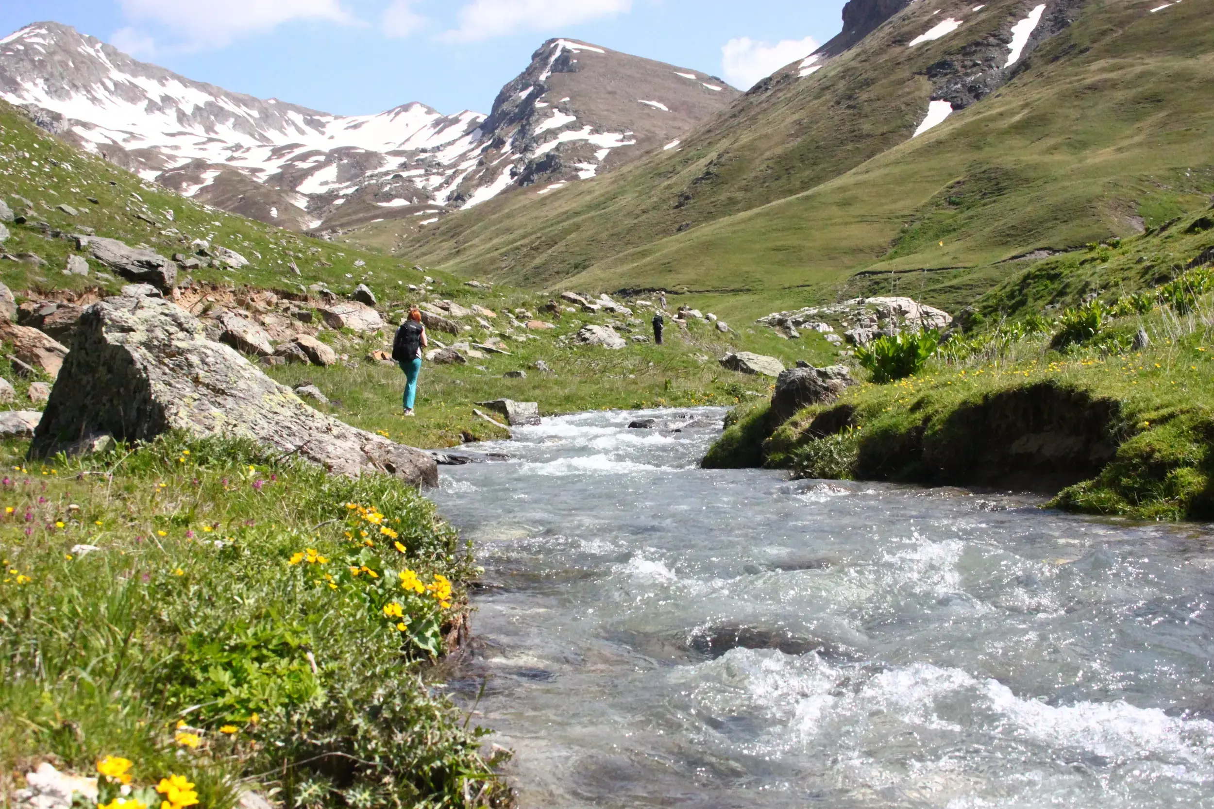 Eine Wanderin pausiert am Ufer eines Gebirgsflusses im Korab-Koritnik Naturpark vor schneebedeckten Gipfeln in Albanien.