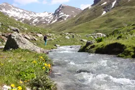 Eine Wanderin pausiert am Ufer eines Gebirgsflusses im Korab-Koritnik Naturpark vor schneebedeckten Gipfeln in Albanien.