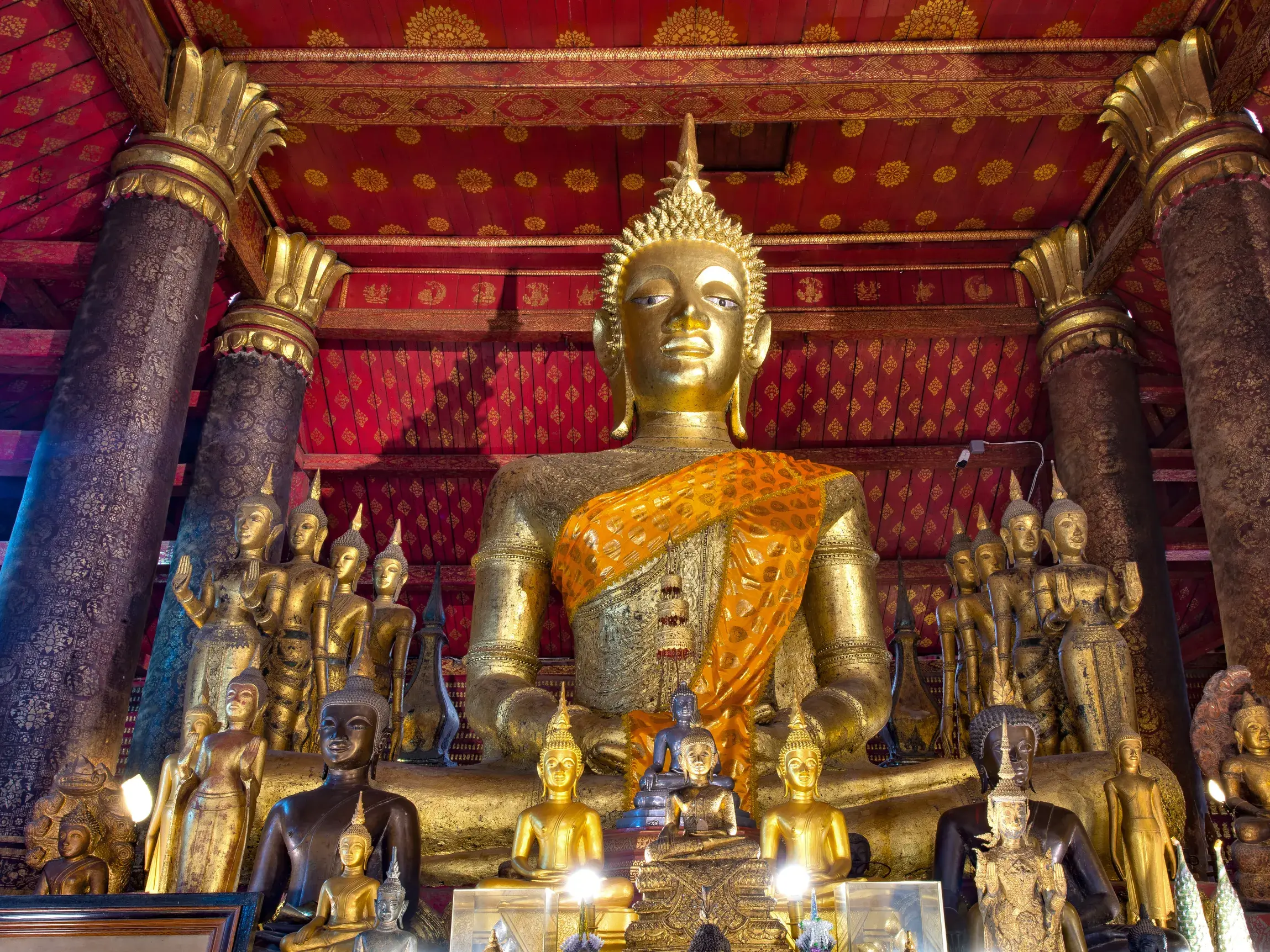 Große goldene Buddha-Statue mit oranger Schärpe im Tempel Wat Mai in Luang Prabang, umgeben von vielen kleineren Figuren.