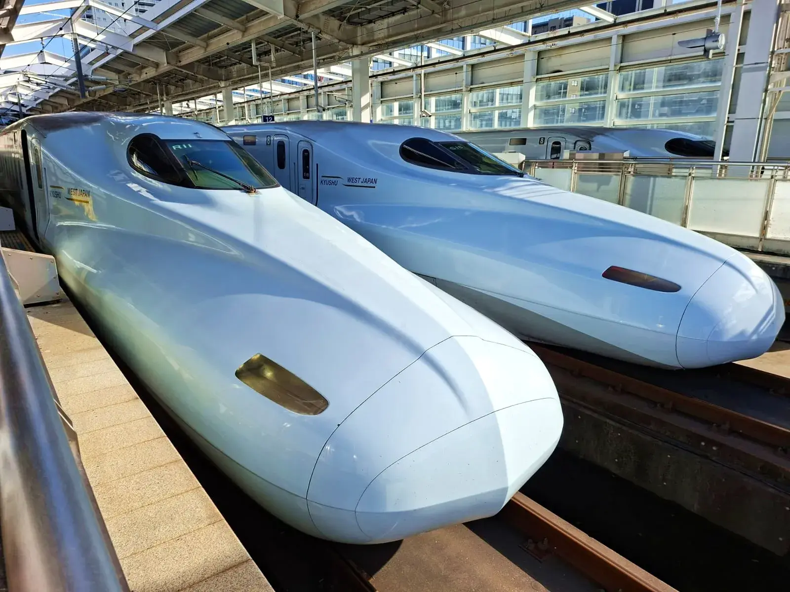 Zwei weiße, aerodynamische Shinkansen-Schnellzüge von West Japan Rail stehen an einem hellen, modernen Bahnhofsbahnsteig.