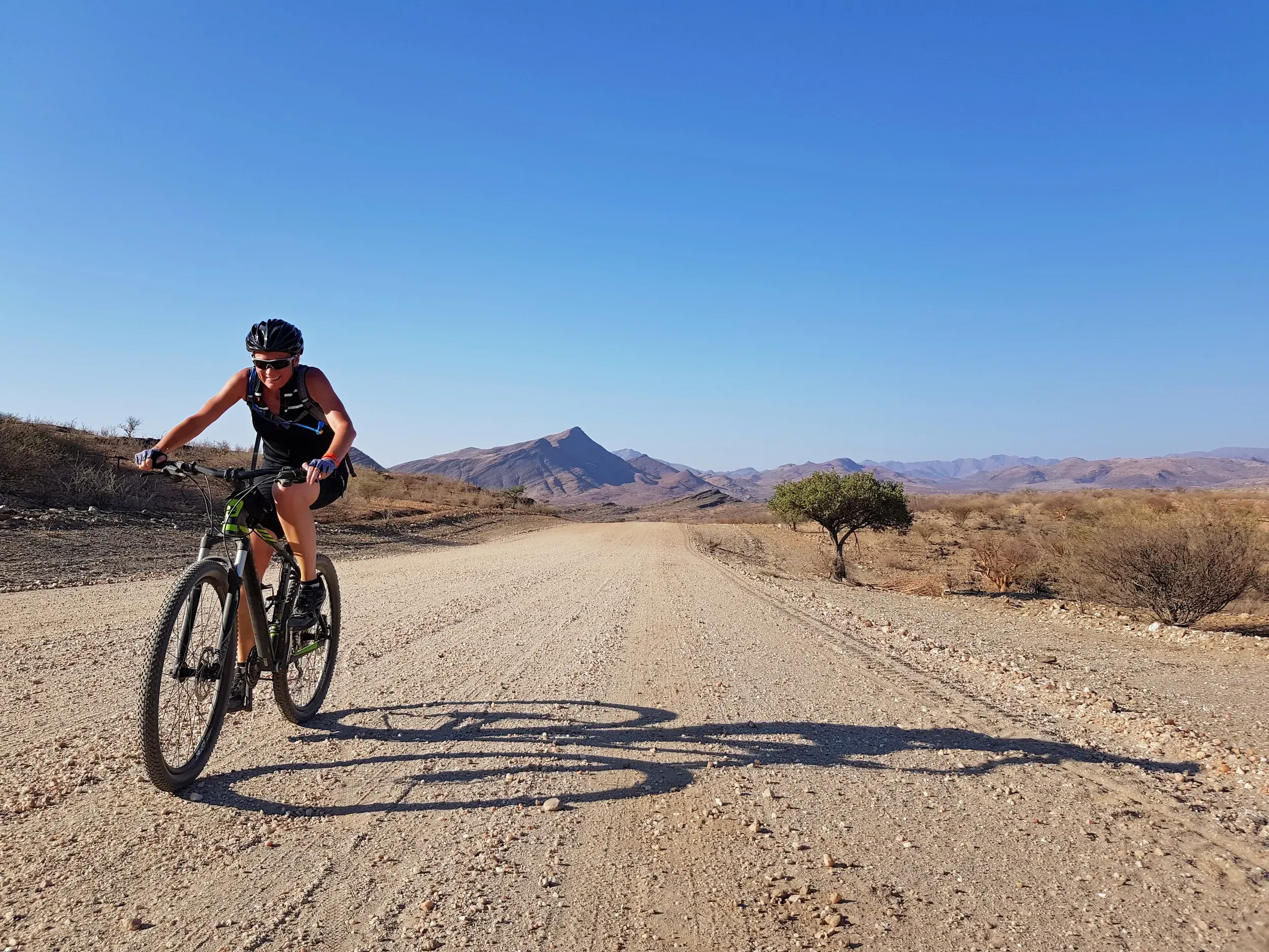 Sonnige Mountainbike-Etappe auf einer Schotterpiste durch die Namib-Wüste mit Blick auf die Bergkette am Horizont.
