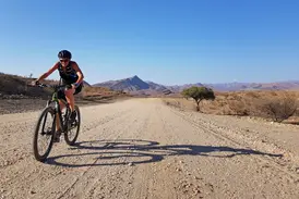 Sonnige Mountainbike-Etappe auf einer Schotterpiste durch die Namib-Wüste mit Blick auf die Bergkette am Horizont.