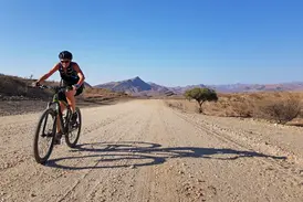 Sonnige Mountainbike-Etappe auf einer Schotterpiste durch die Namib-Wüste mit Blick auf die Bergkette am Horizont.