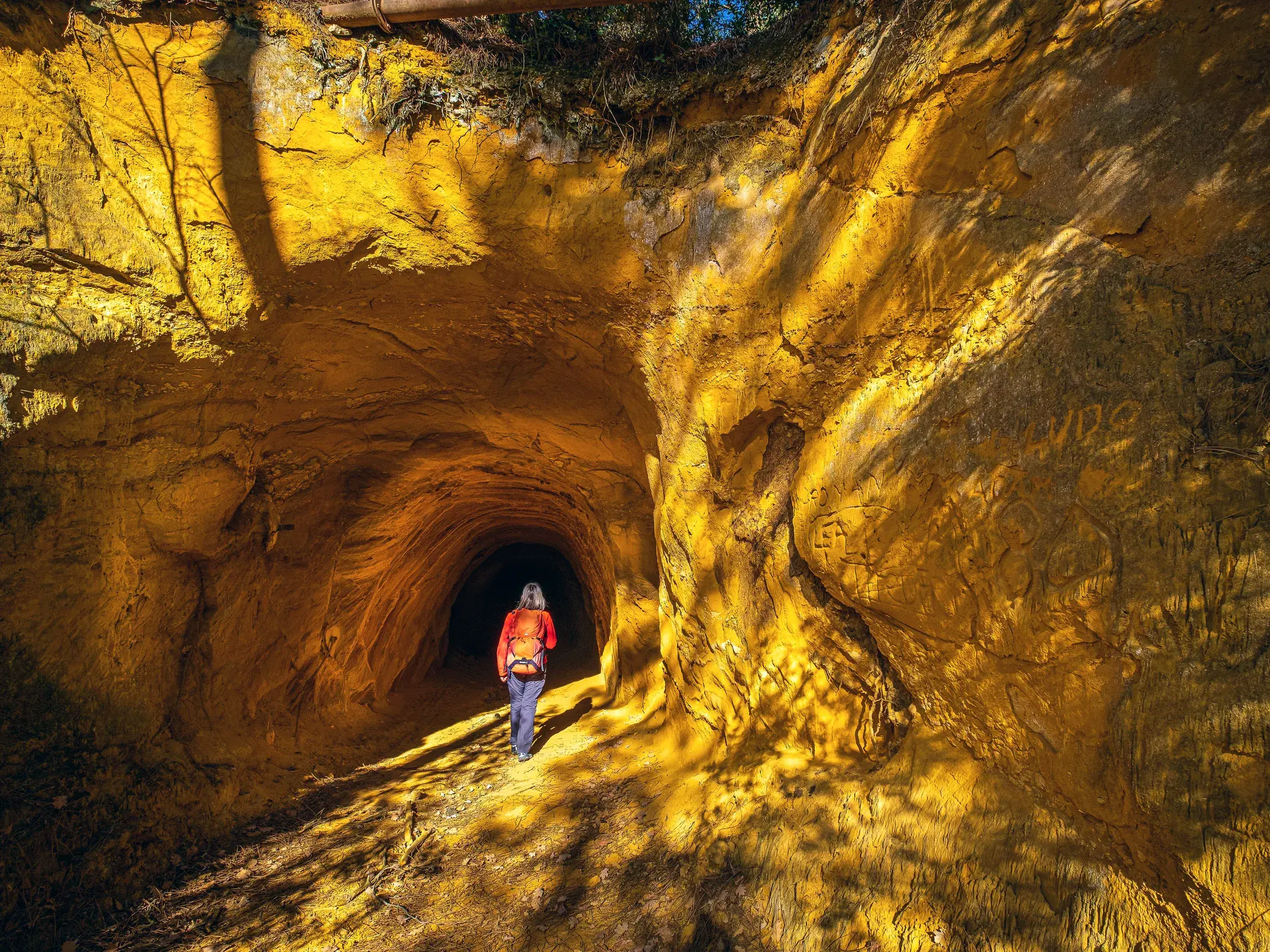Reisende mit Rucksack geht durch einen ockergelben Felstunnel in das Innere einer Höhle.