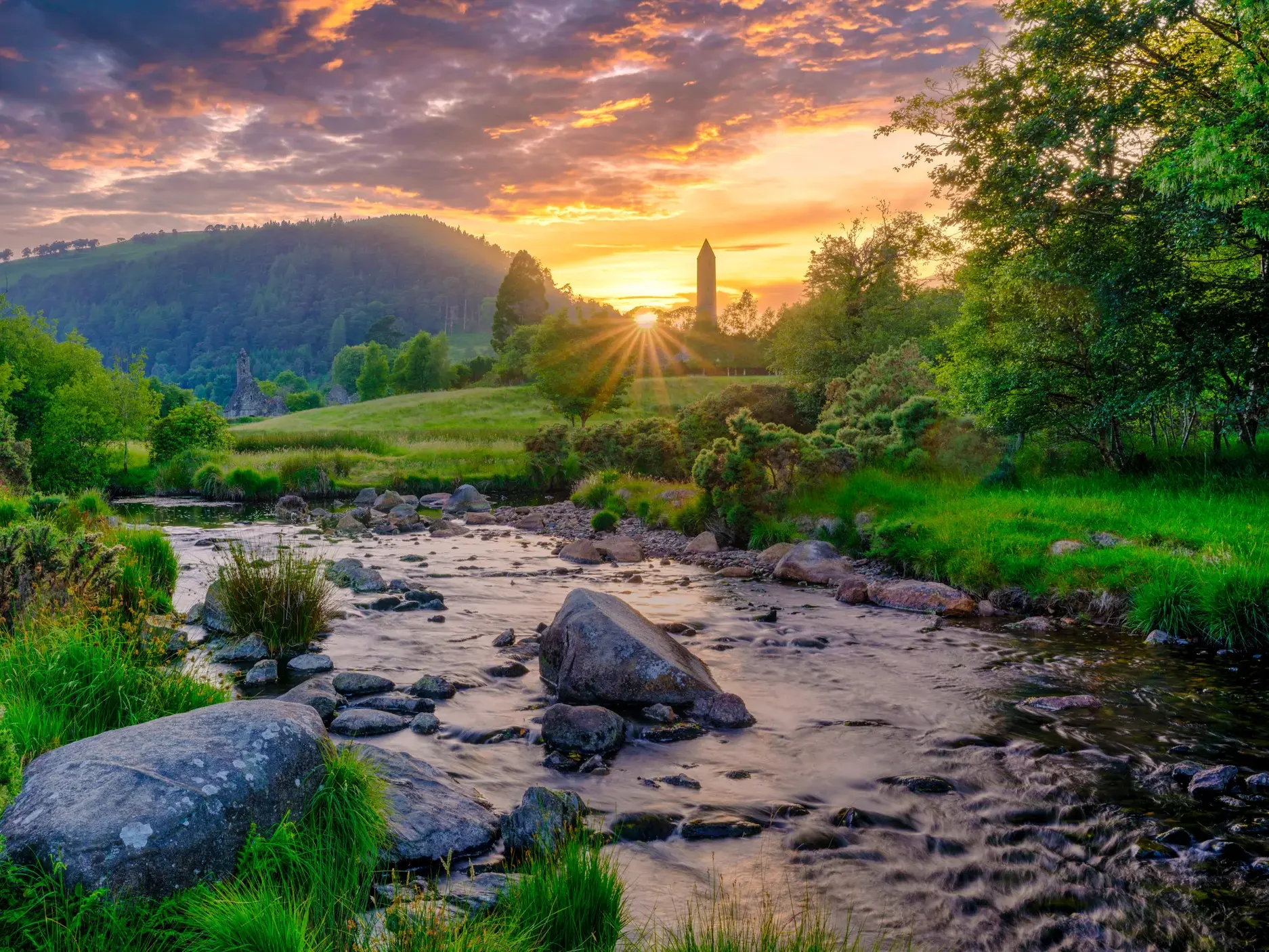Ein Bach fließt durch das grüne Tal von Glendalough in den Wicklowbergen, im Hintergrund der Rundturm der Klosteranlage bei Sonnenuntergang.