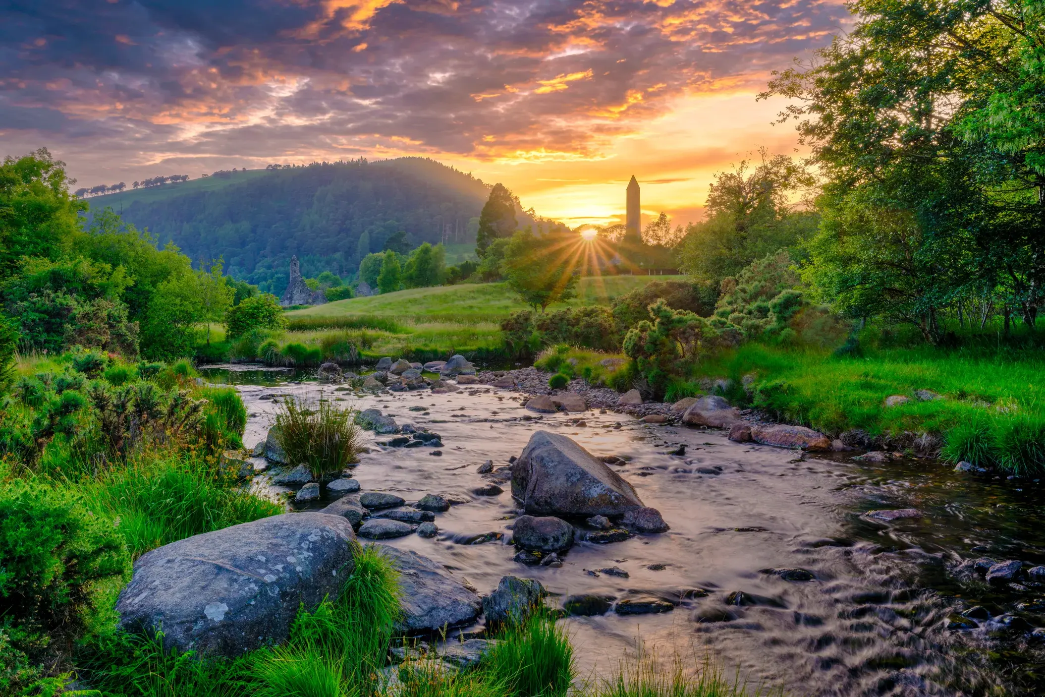 Ein Bach fließt durch das grüne Tal von Glendalough in den Wicklowbergen, im Hintergrund der Rundturm der Klosteranlage bei Sonnenuntergang.