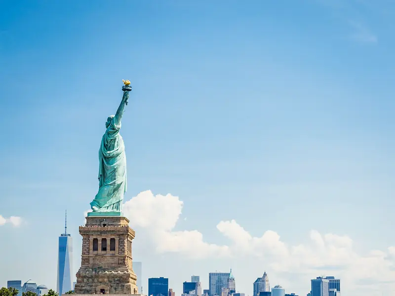 Blick vom Wasser auf die Freiheitsstatue auf Liberty Island mit der Skyline von Lower Manhattan und dem One World Trade Center.