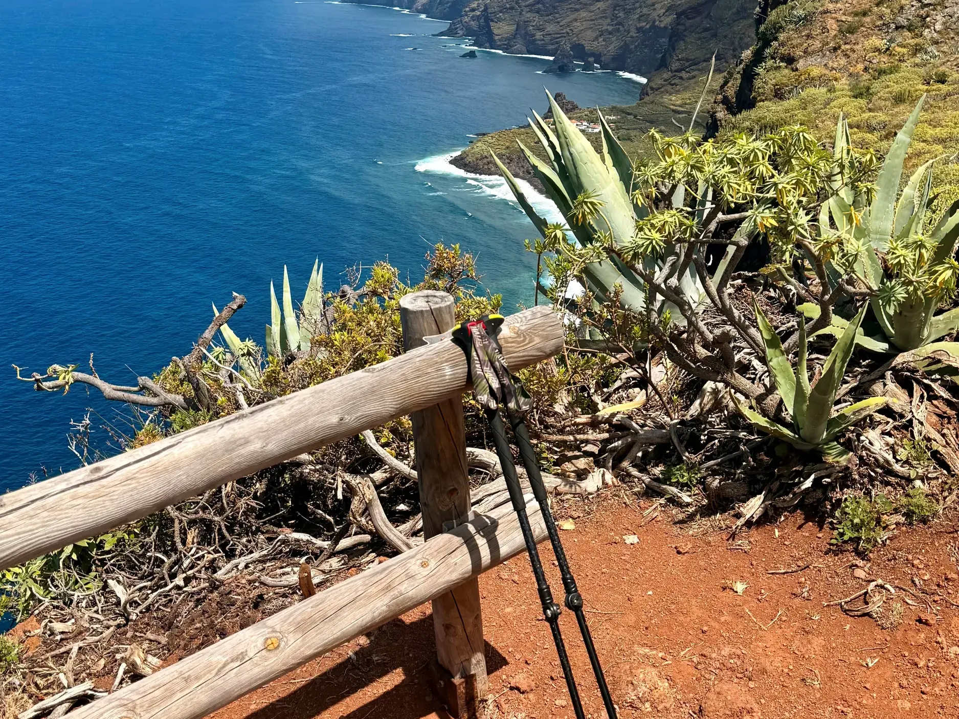 Trekkingstöcke lehnen an einem Holzgeländer mit Blick auf die steile, grüne Küste und den tiefblauen Atlantik auf La Palma.