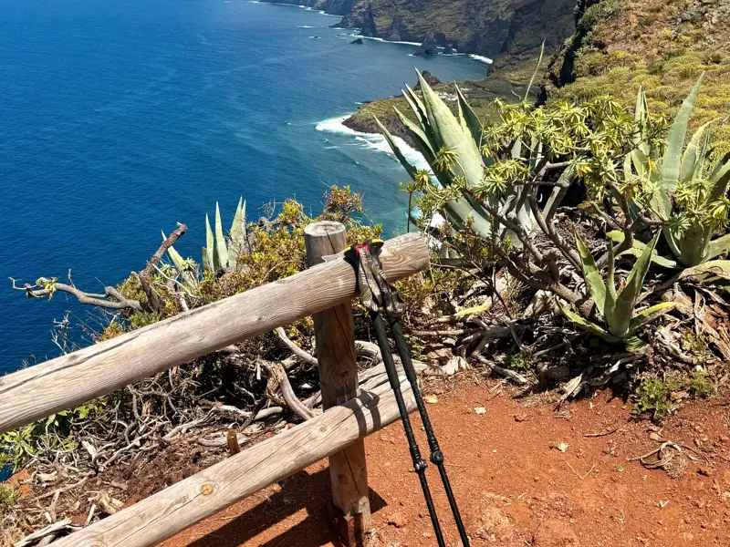 Trekkingstöcke lehnen an einem Holzgeländer mit Blick auf die steile, grüne Küste und den tiefblauen Atlantik auf La Palma.
