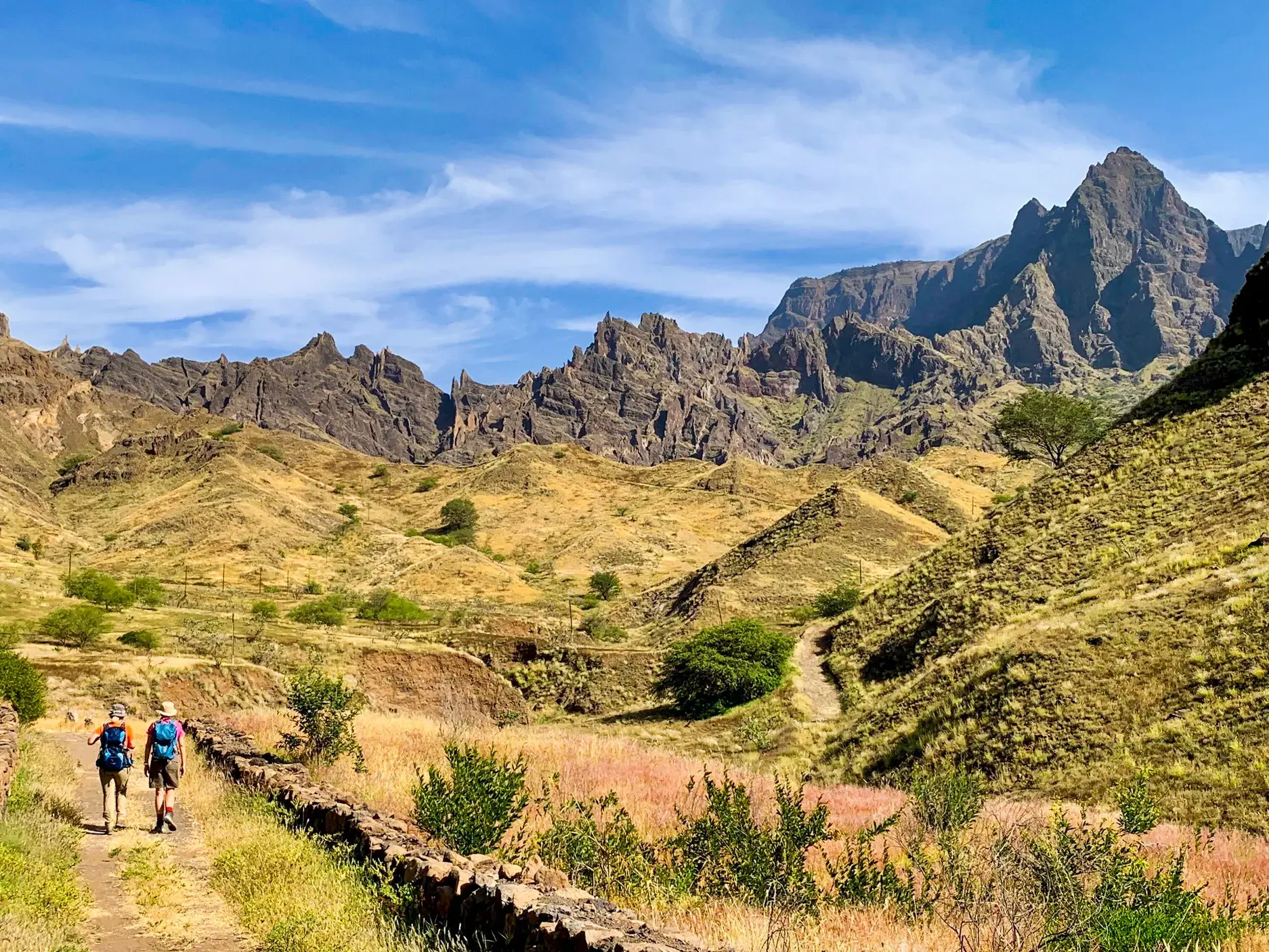 Zwei Trekker auf einem Steinpfad durch eine karge Hochebene auf Santo Antão vor einer schroffen Bergkette auf den Kapverden.