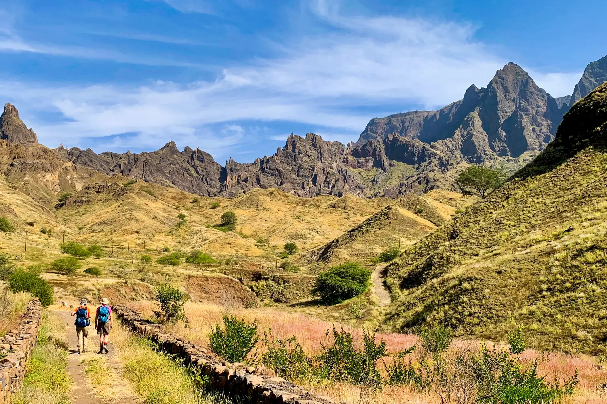 Zwei Trekker auf einem Steinpfad durch eine karge Hochebene auf Santo Antão vor einer schroffen Bergkette auf den Kapverden.