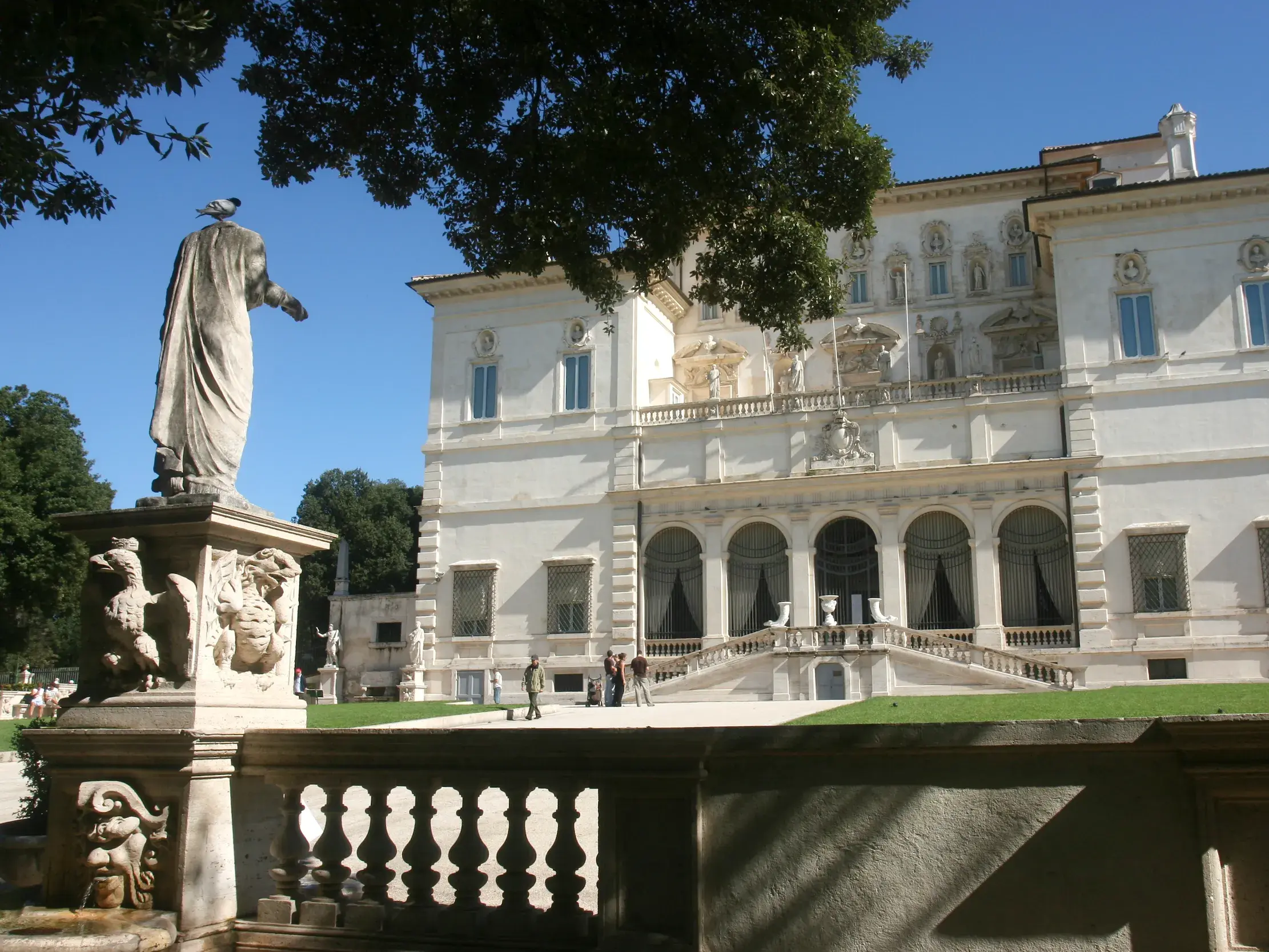 Die prunkvolle, weiße Fassade der Galleria Borghese in Rom, umgeben von ihrem Park mit einer klassischen Statue im Vordergrund.