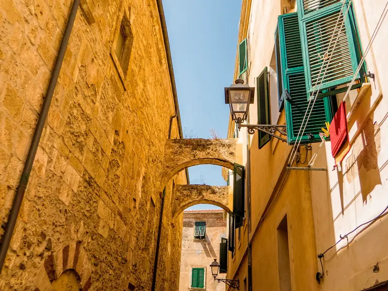 Blick durch eine enge Gasse mit sonnenbeschienenen, gelben Steinfassaden und Steinbögen in der Altstadt von Alghero.