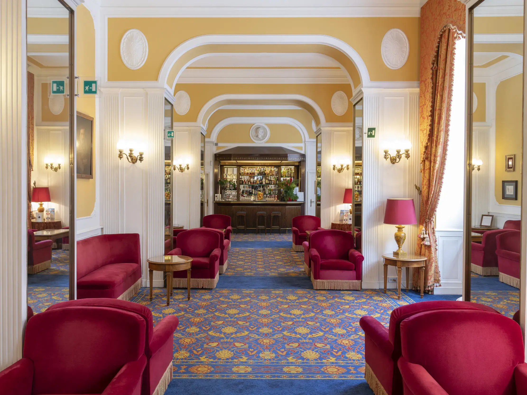 Die elegante Lobby des Hotel Massimo d'Azeglio in Rom mit roten Samtsesseln, blauem Teppich und der Hotelbar im Hintergrund.