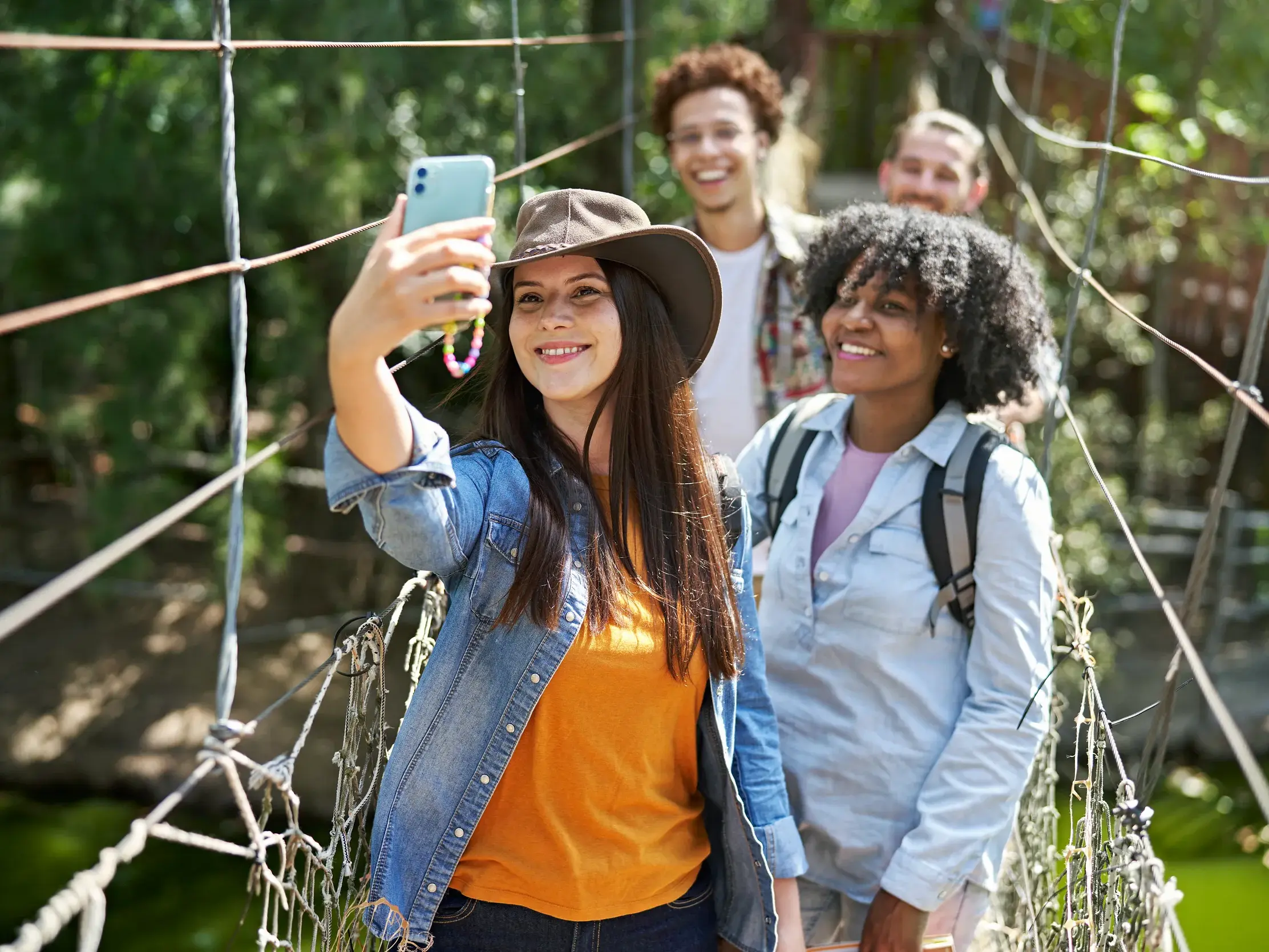 Eine junge Frau mit Hut macht ein fröhliches Selfie mit ihrer Reisegruppe auf einer Seilbrücke im Lake-Manyara-Nationalpark.
