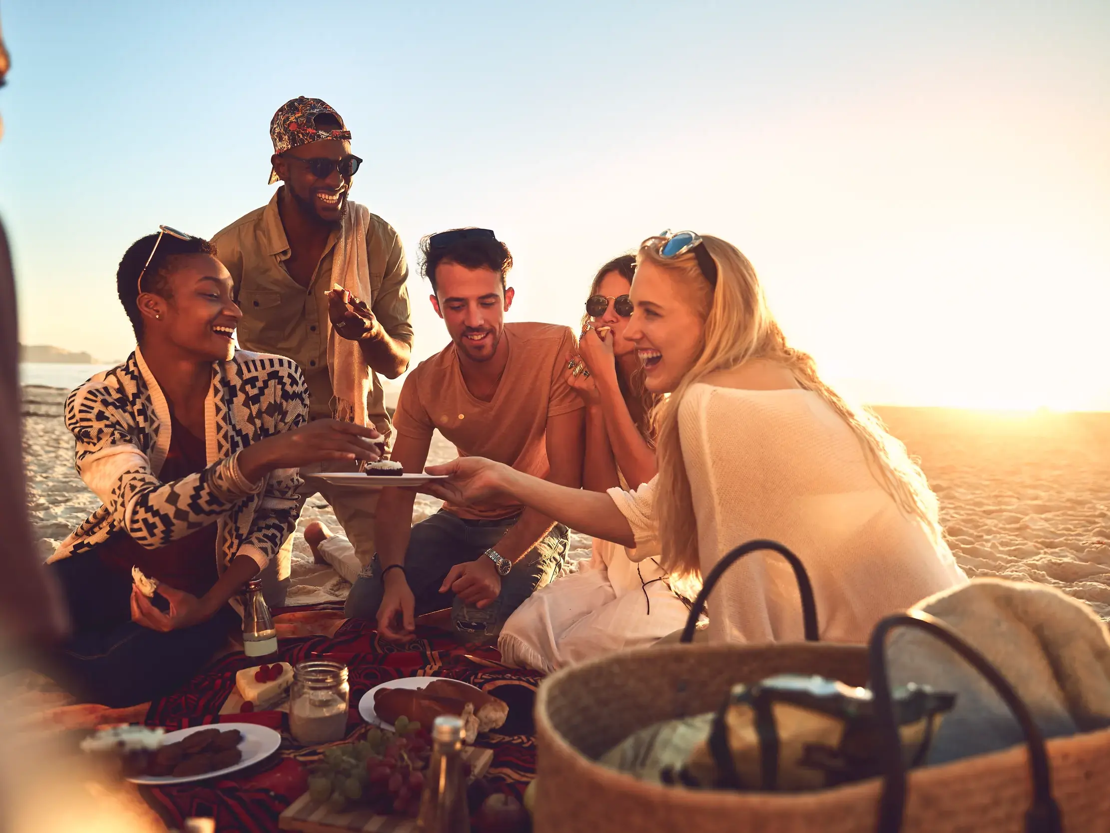 Junge Reisende teilen lachend Essen bei einem Picknick am Sandstrand der Baja California bei Sonnenuntergang.