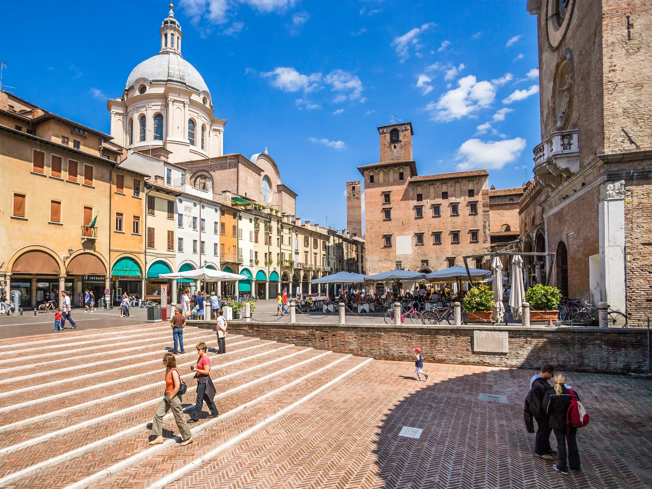 Besucher flanieren über die sonnige Piazza delle Erbe in Mantua mit Blick auf die Fassaden und die Kuppel der Basilica di Sant'Andrea.