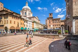 Besucher flanieren über die sonnige Piazza delle Erbe in Mantua mit Blick auf die Fassaden und die Kuppel der Basilica di Sant'Andrea.
