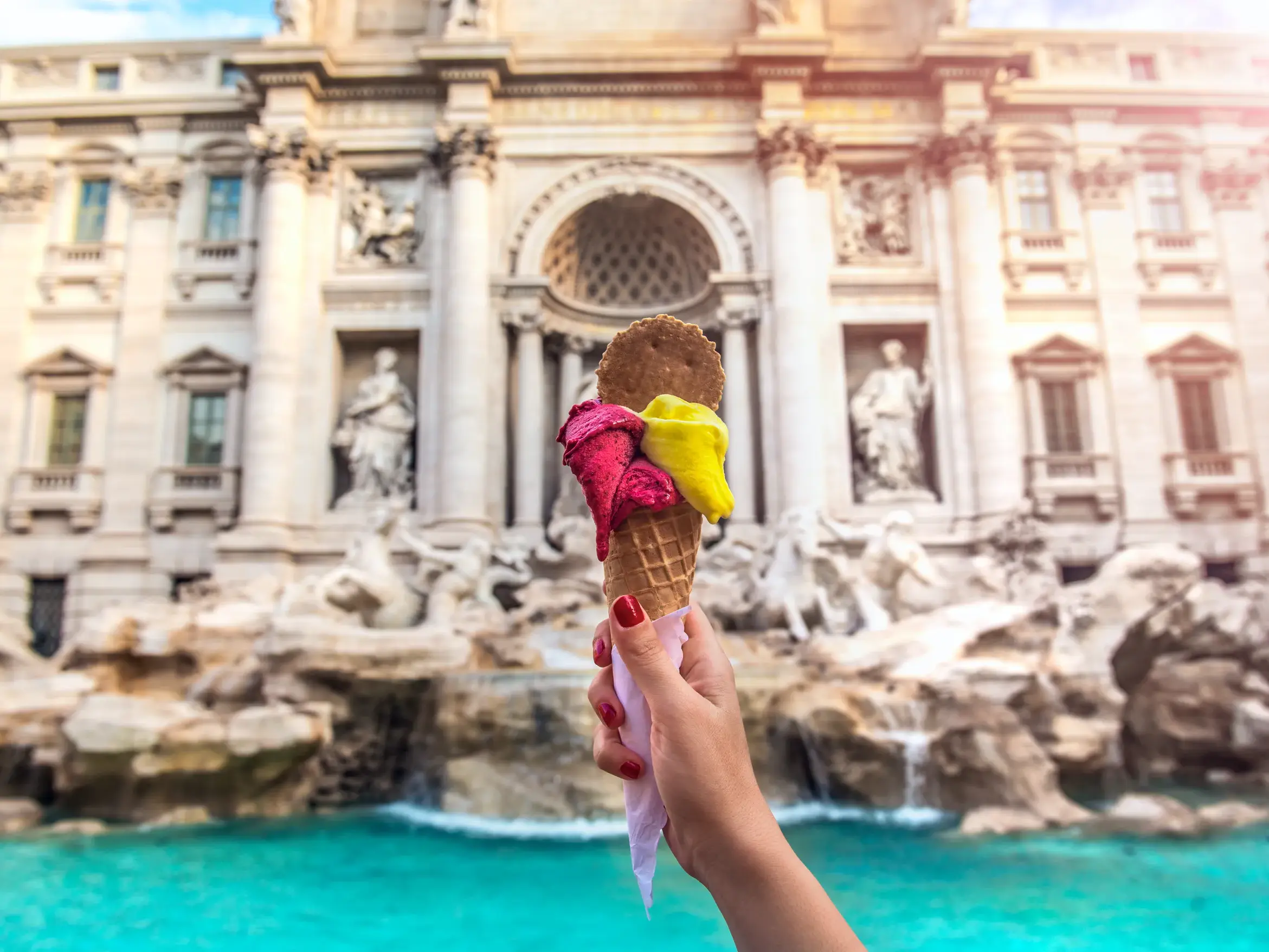 Frauenhand hält eine Eistüte mit buntem Gelato vor den berühmten Trevi-Brunnen in Rom, Sinnbild des Dolce Vita.
