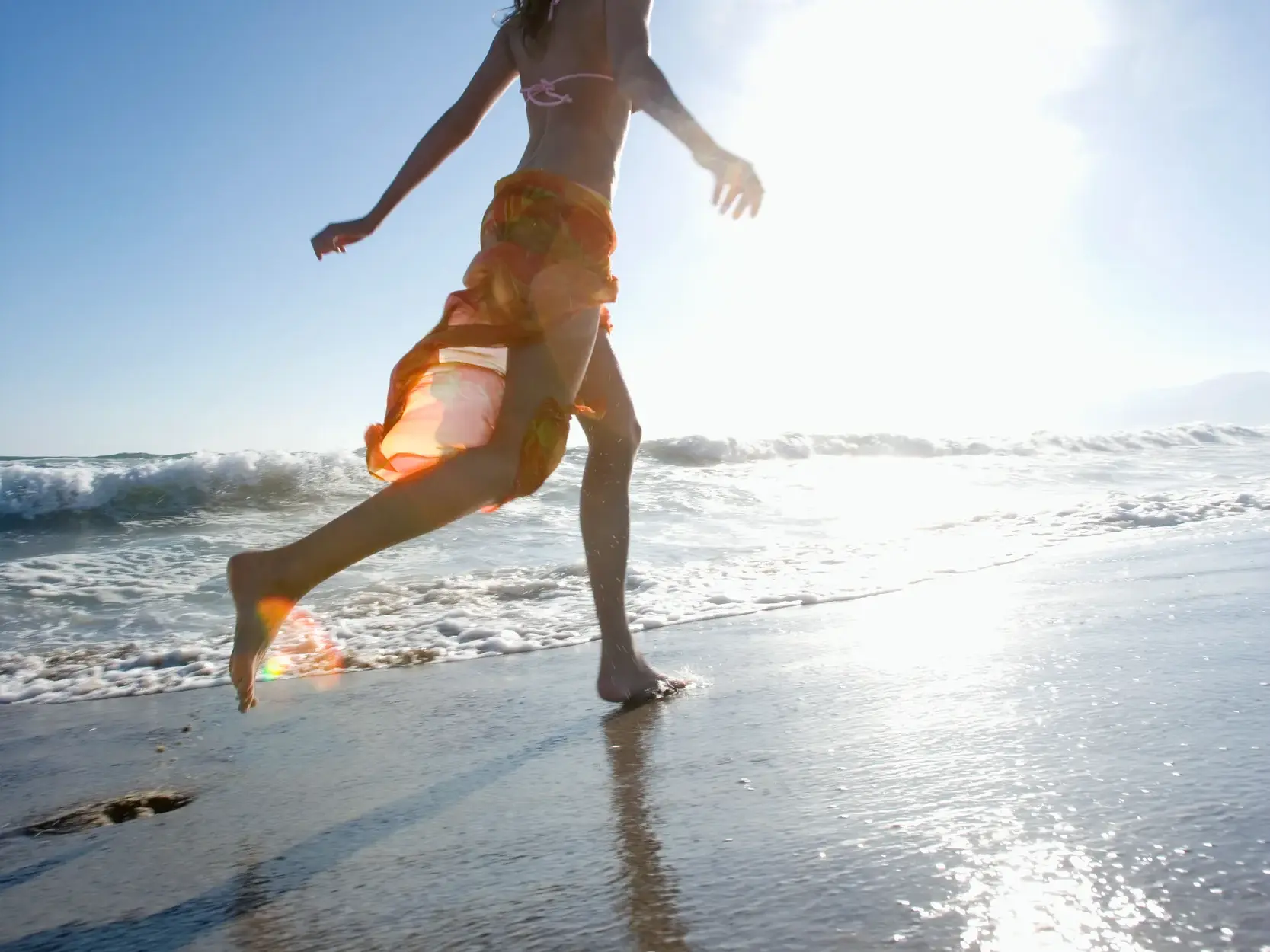 Im hellen Sonnenlicht läuft eine Frau mit einem Pareo bekleidet am Strand von Numana durch die seichten Wellen der Adria.