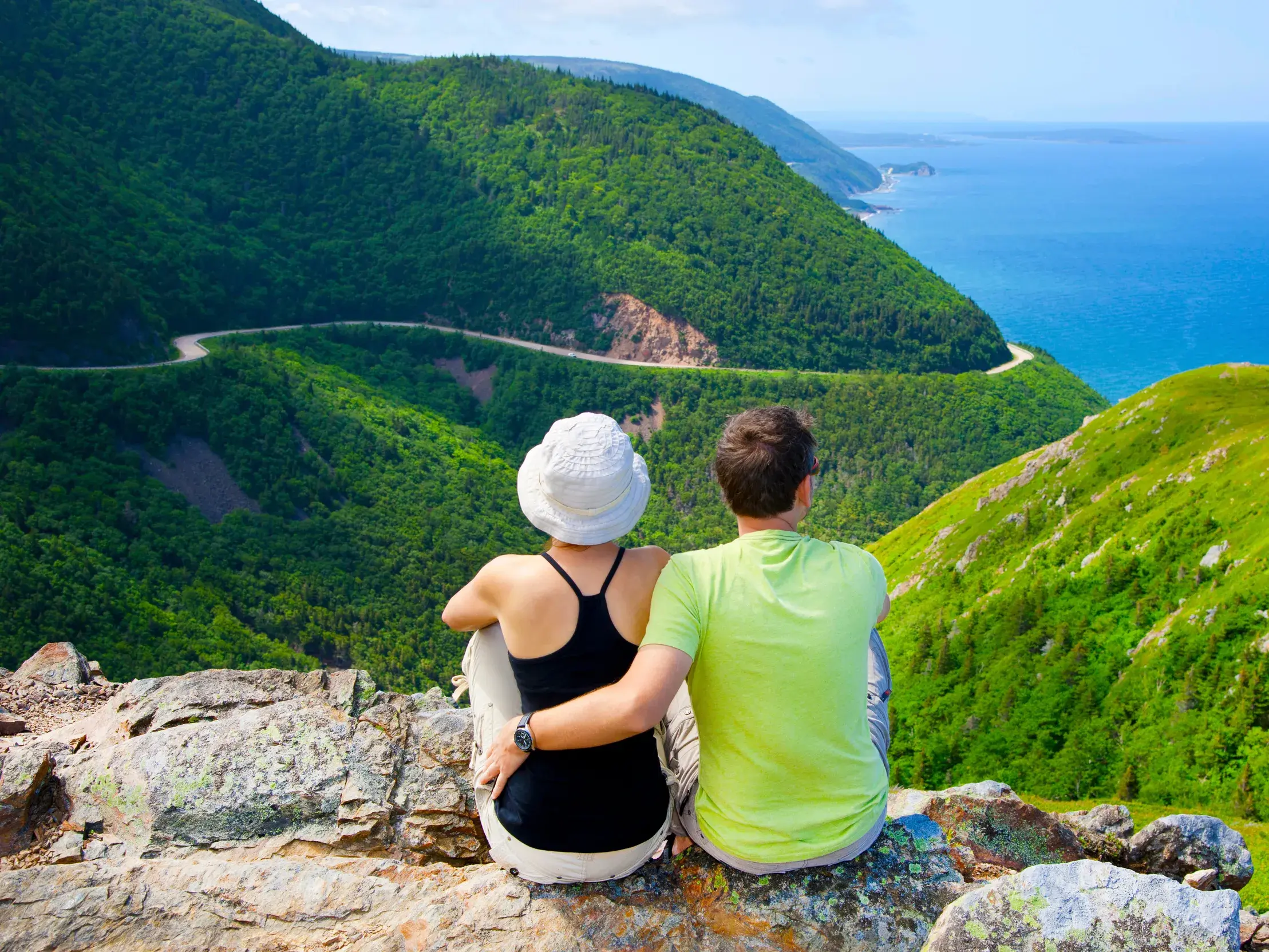 Zwei Wanderer blicken von einem Felsen auf den kurvigen Cabot Trail und die Küste im Cape Breton Highlands Nationalpark.