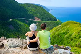 Zwei Wanderer blicken von einem Felsen auf den kurvigen Cabot Trail und die Küste im Cape Breton Highlands Nationalpark.