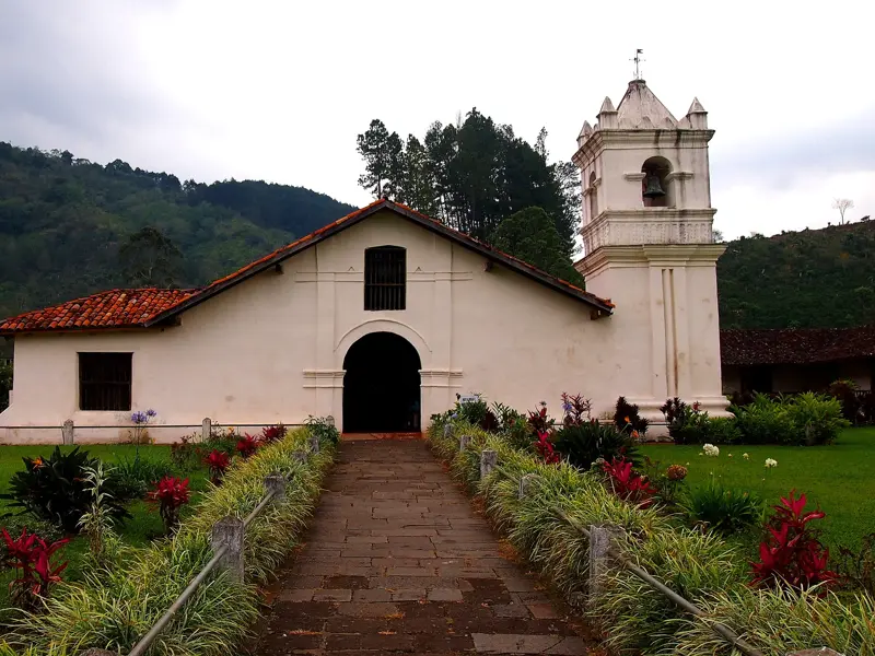 Ein Steinweg führt zur historischen, weißen Kirche von Orosi, umgeben von bunten Blumen und den grünen Hügeln Costa Ricas.
