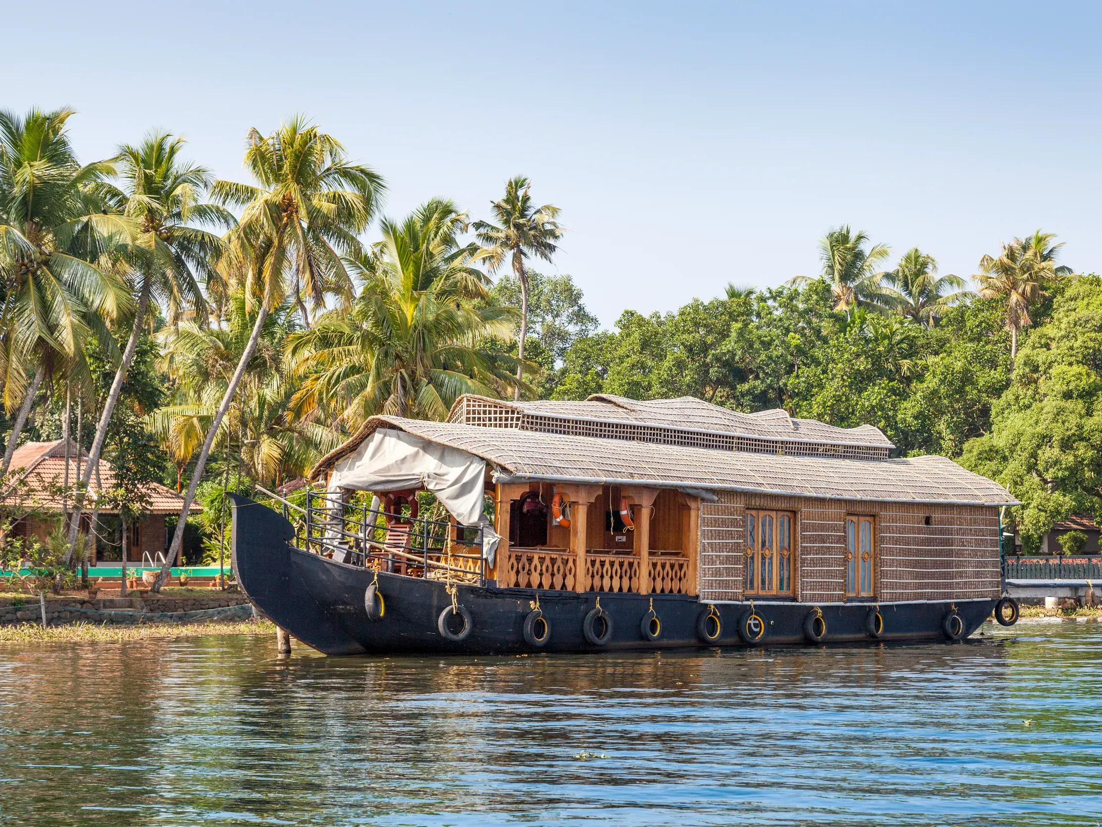 Ein traditionelles Hausboot ankert in der Lagunenlandschaft der Kerala Backwaters in Südindien, im Hintergrund ragen hohe Palmen auf.