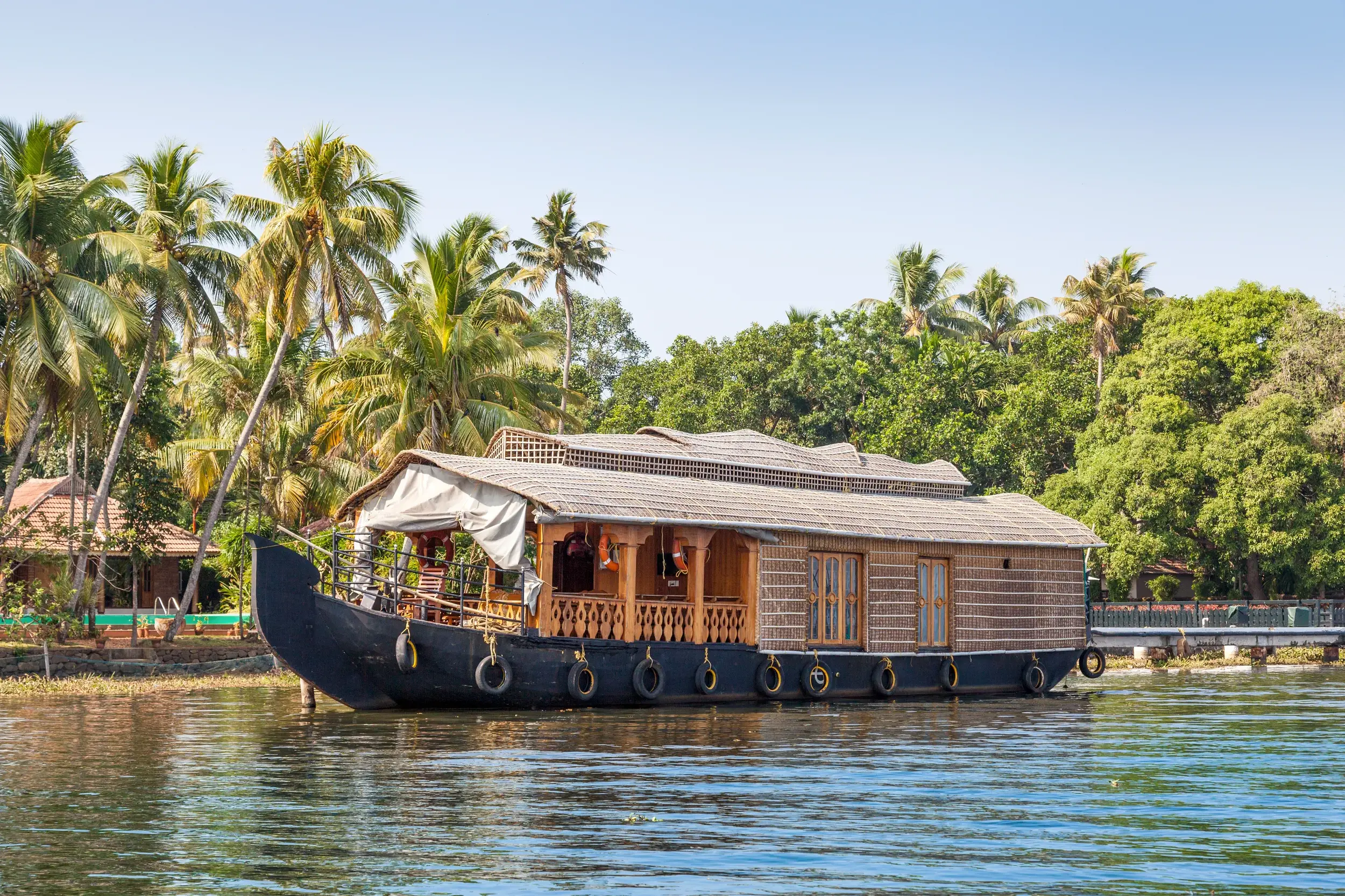 Ein traditionelles Hausboot ankert in der Lagunenlandschaft der Kerala Backwaters in Südindien, im Hintergrund ragen hohe Palmen auf.
