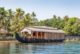 Ein traditionelles Hausboot ankert in der Lagunenlandschaft der Kerala Backwaters in Südindien, im Hintergrund ragen hohe Palmen auf.