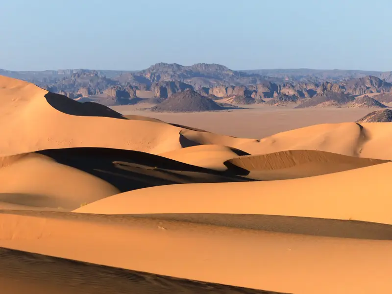 Sanft geschwungene, orangefarbene Dünen des Erg Admer in der Sahara vor der Felskulisse des Tassili n'Ajjer Plateaus.