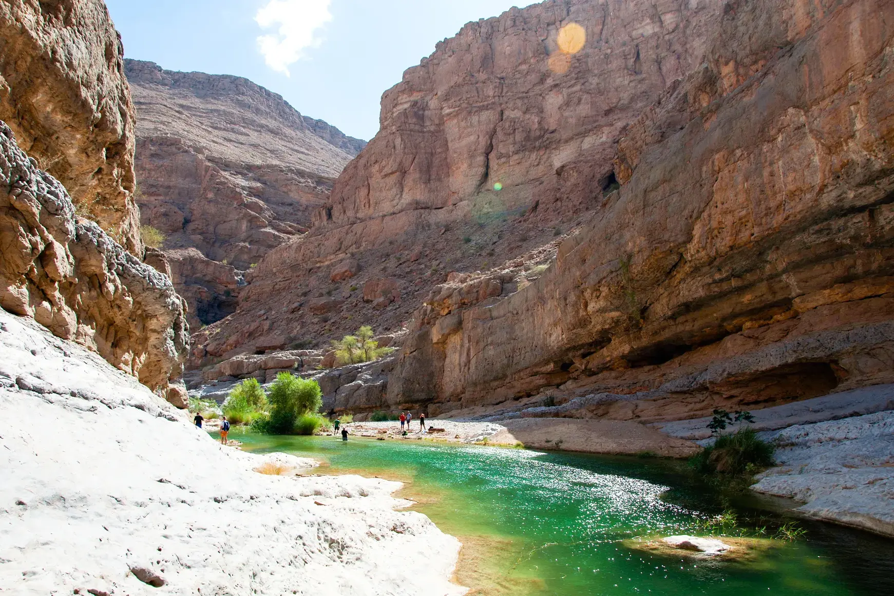 Eine Wandergruppe erkundet das Ufer eines türkisfarbenen Flusses, der sich durch die Felsenschlucht des Wadi Shab im Oman windet.