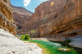 Eine Wandergruppe erkundet das Ufer eines türkisfarbenen Flusses, der sich durch die Felsenschlucht des Wadi Shab im Oman windet.