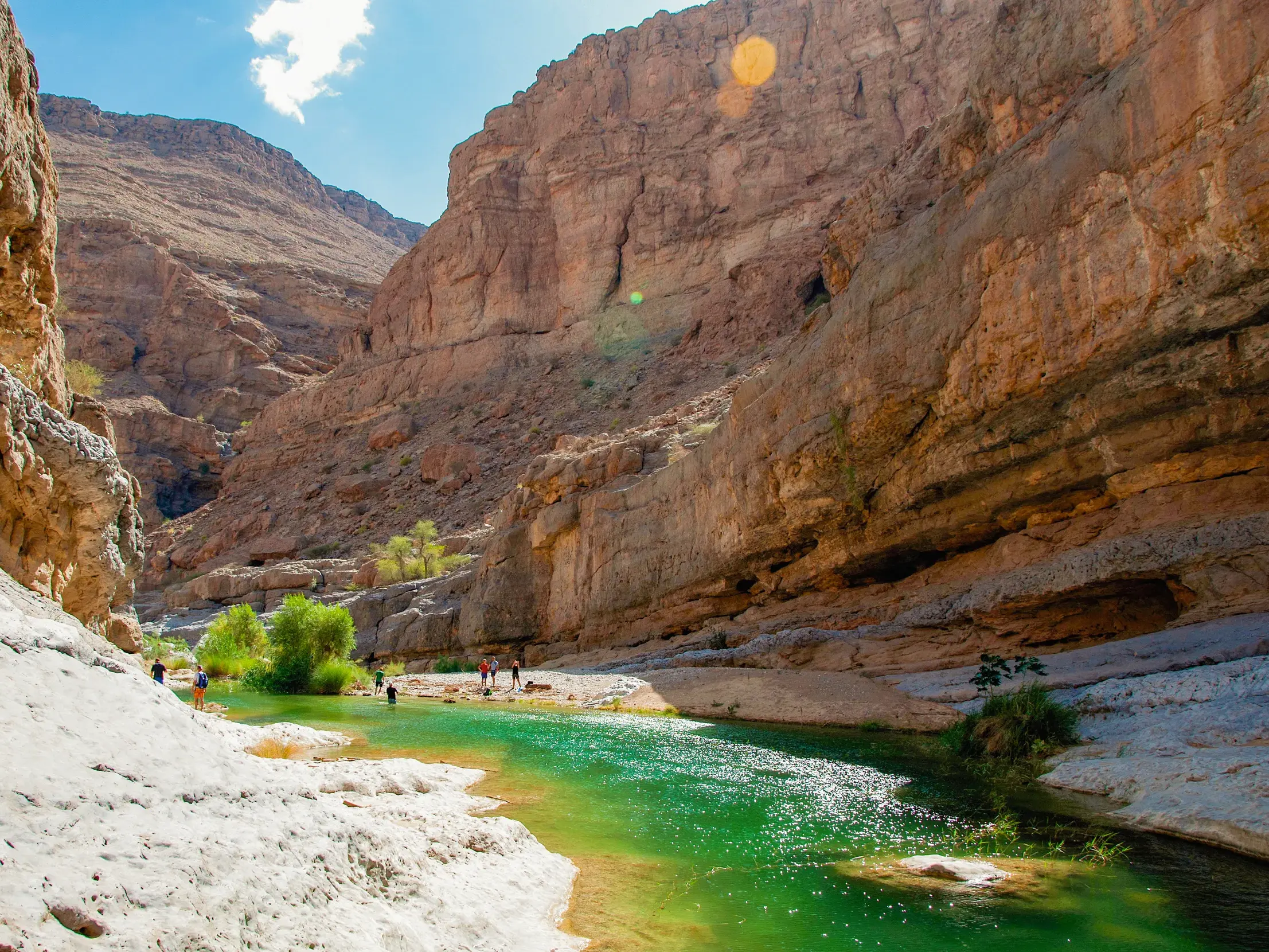 Eine Wandergruppe erkundet ein Wadi im Oman mit türkisfarbenen Wasserbecken umgeben von steilen, sonnigen Felswänden.