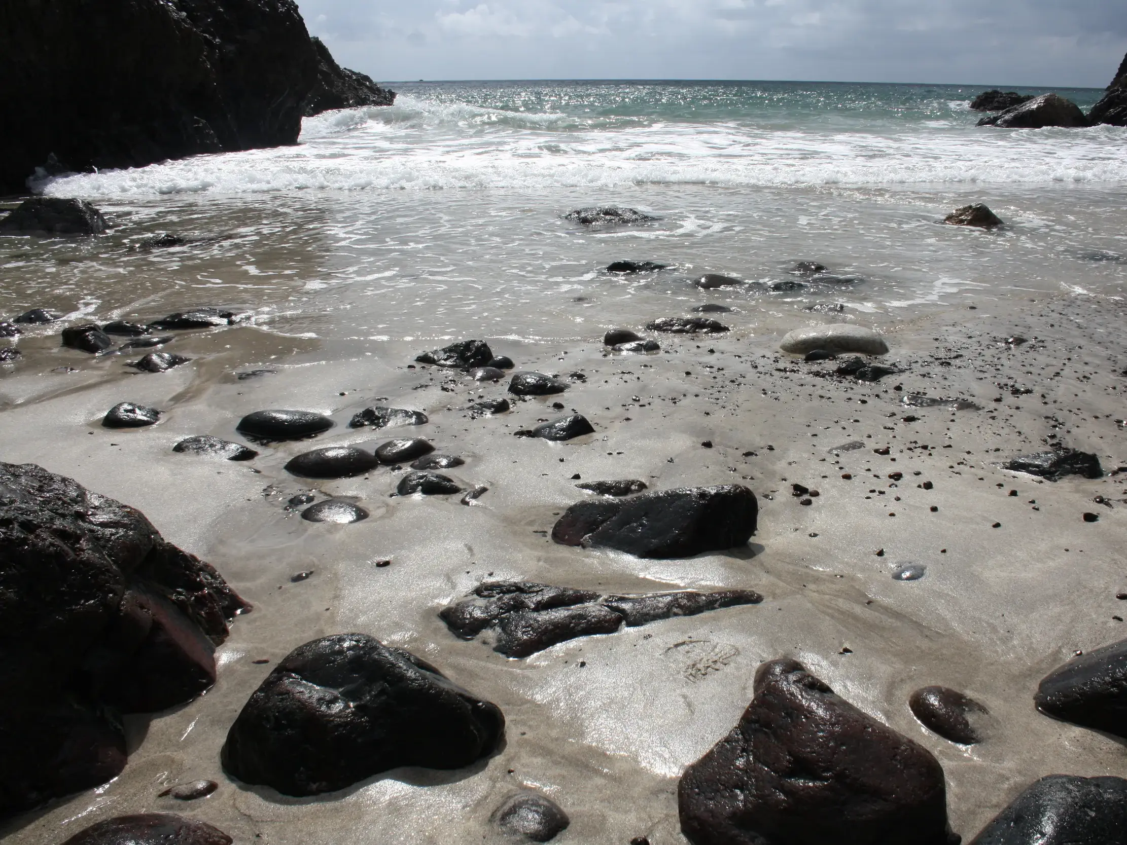 Nasse Felsen am Sandstrand einer einsamen Bucht am South West Coast Path, wo Wellen sanft an die Küste Cornwalls rollen.