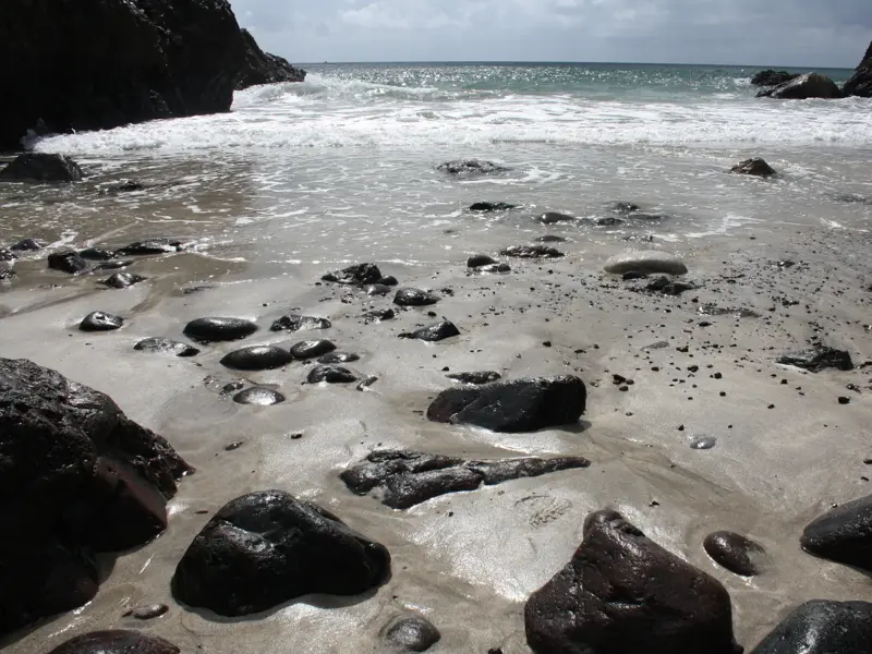 Nasse Felsen am Sandstrand einer einsamen Bucht am South West Coast Path, wo Wellen sanft an die Küste Cornwalls rollen.