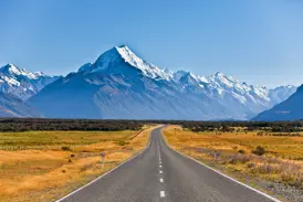 Eine leere Asphaltstraße führt durch goldenes Grasland auf den majestätischen, schneebedeckten Aoraki / Mount Cook zu.