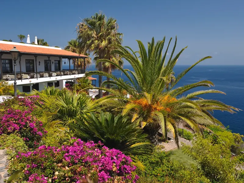 Das Hotel Jardín Tecina auf La Gomera mit üppigem Garten, Palmen und Blick auf den tiefblauen Atlantik bei Sonnenschein.