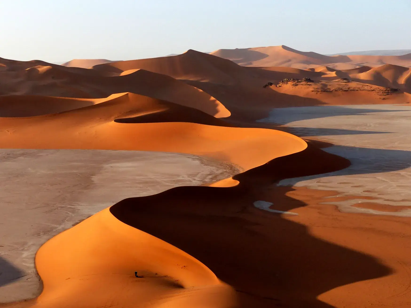 Ein einzelner Wanderer am Fuße einer riesigen, orangeroten Düne in der weiten Wüstenlandschaft des Tadrart in Algerien.