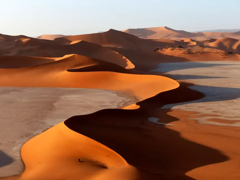 Ein einzelner Wanderer am Fuße einer riesigen, orangeroten Düne in der weiten Wüstenlandschaft des Tadrart in Algerien.