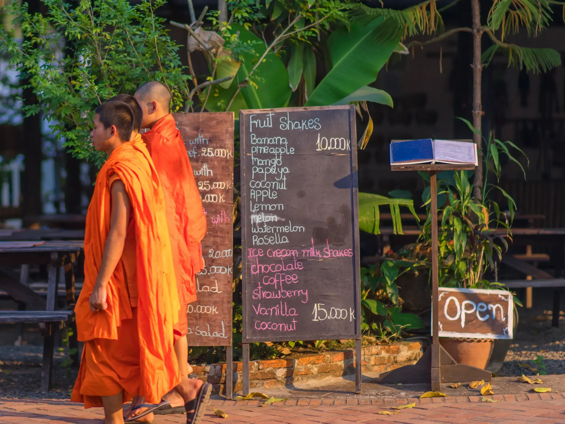 Zwei buddhistische Mönche in orangefarbenen Roben gehen in Luang Prabang, Laos, an einem Café mit Kreidetafel-Menü vorbei.