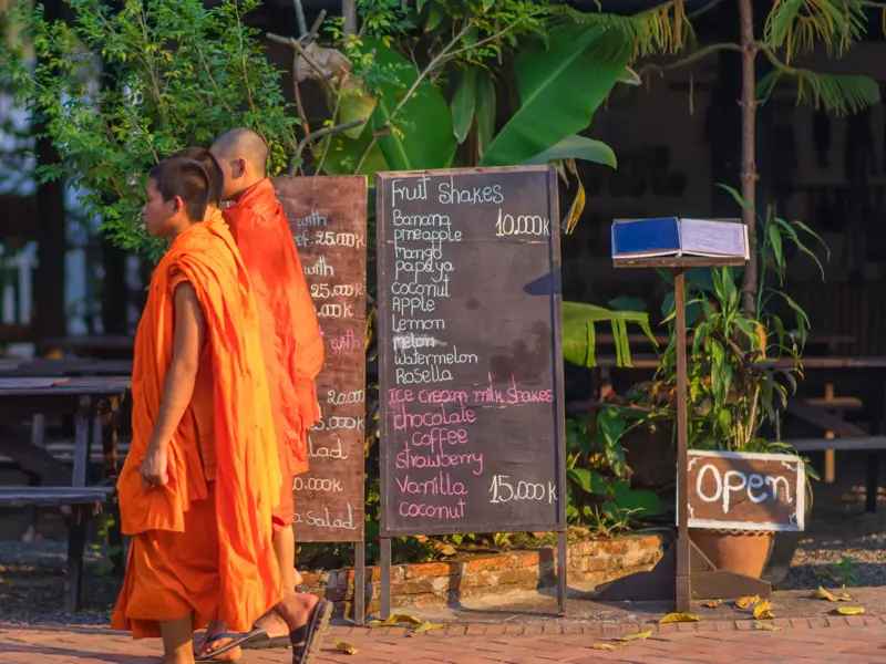 Zwei buddhistische Mönche in orangefarbenen Roben gehen in Luang Prabang, Laos, an einem Café mit Kreidetafel-Menü vorbei.