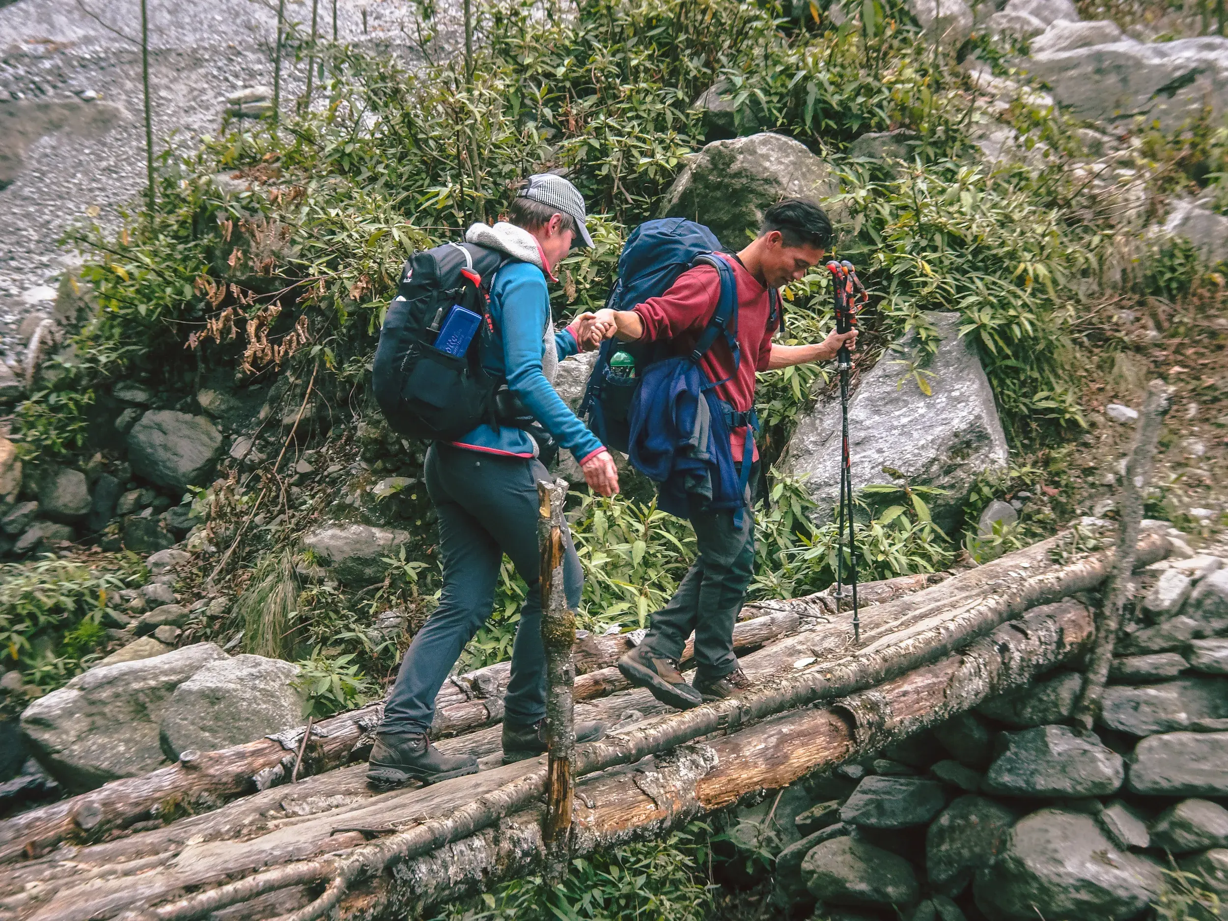 Ein nepalesischer Reiseleiter hilft einer Wanderin mit Rucksack beim Trekking in Nepal sicher über eine Holzbrücke.