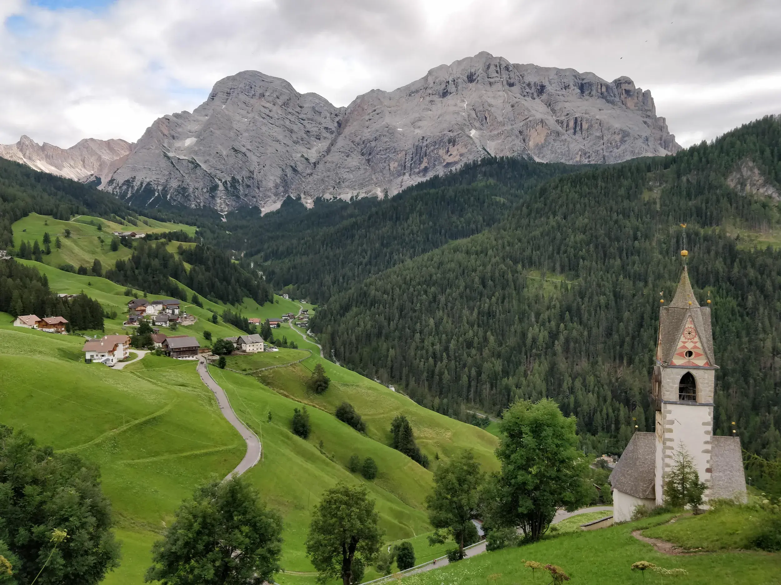 Die St. Barbara Kirche in La Valle (Wengen) inmitten grüner Almwiesen vor der zerklüfteten Felsenkulisse der Dolomiten in Südtirol.