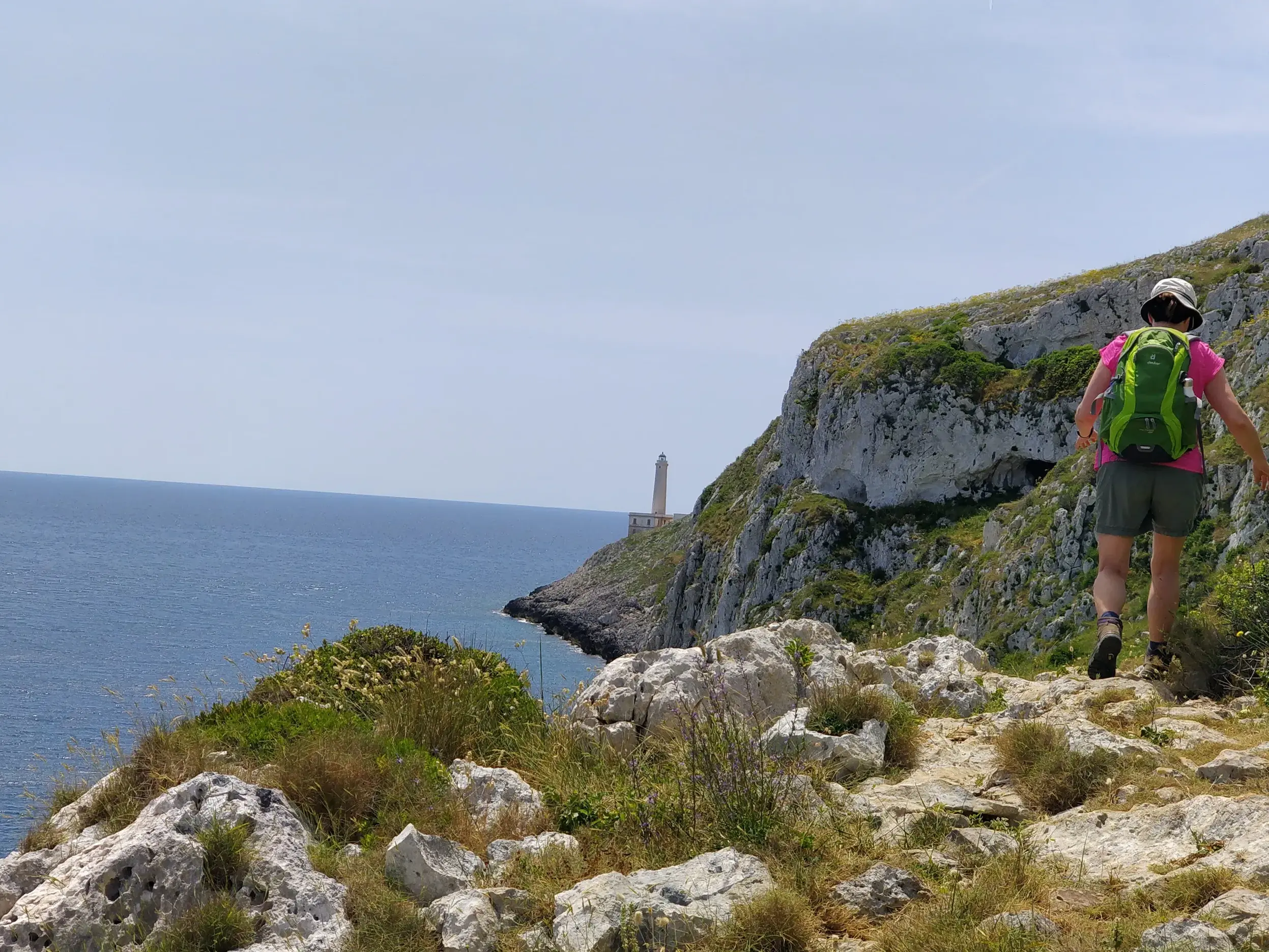 Trekking in Apulien: Wanderin auf einem felsigen Küstenpfad mit Blick auf den Leuchtturm von Punta Palascia und das Meer.