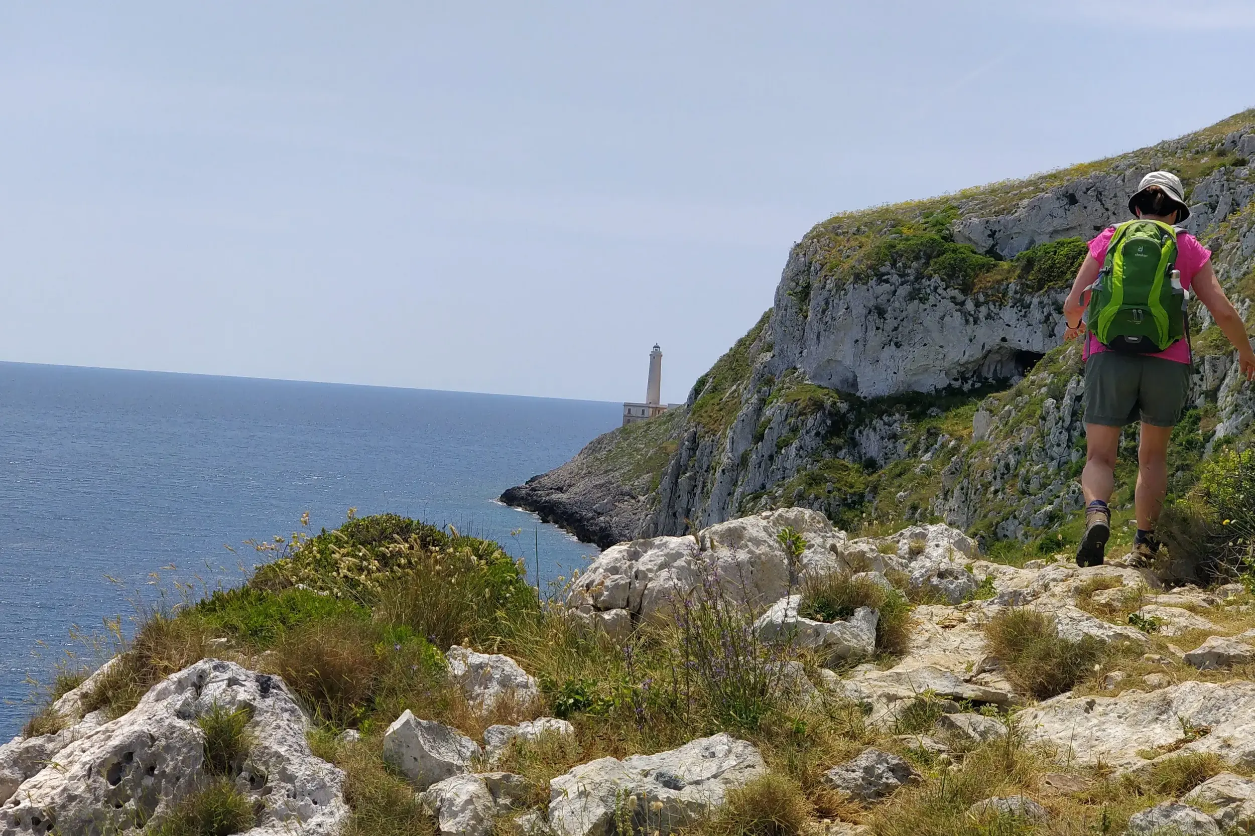 Trekking in Apulien: Wanderin auf einem felsigen Küstenpfad mit Blick auf den Leuchtturm von Punta Palascia und das Meer.