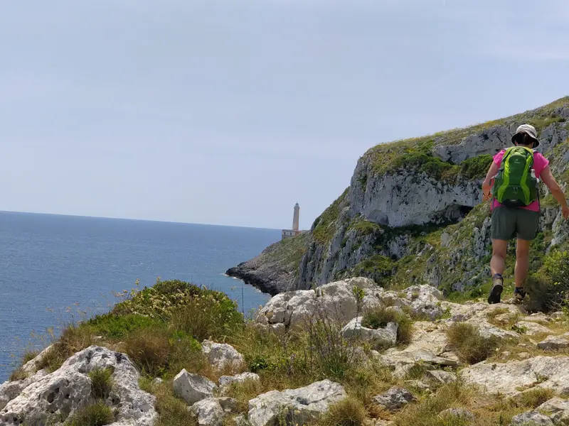 Trekking in Apulien: Wanderin auf einem felsigen Küstenpfad mit Blick auf den Leuchtturm von Punta Palascia und das Meer.
