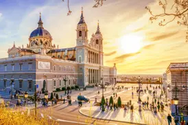 Menschen flanieren vor der Almudena-Kathedrale in Madrid, die von der Abendsonne in goldenes Licht getaucht wird.